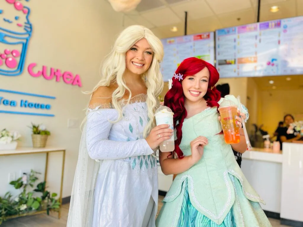 Two costumed characters, Elsa & Ariel, hold drinks inside a CUTEA bubble tea shop.