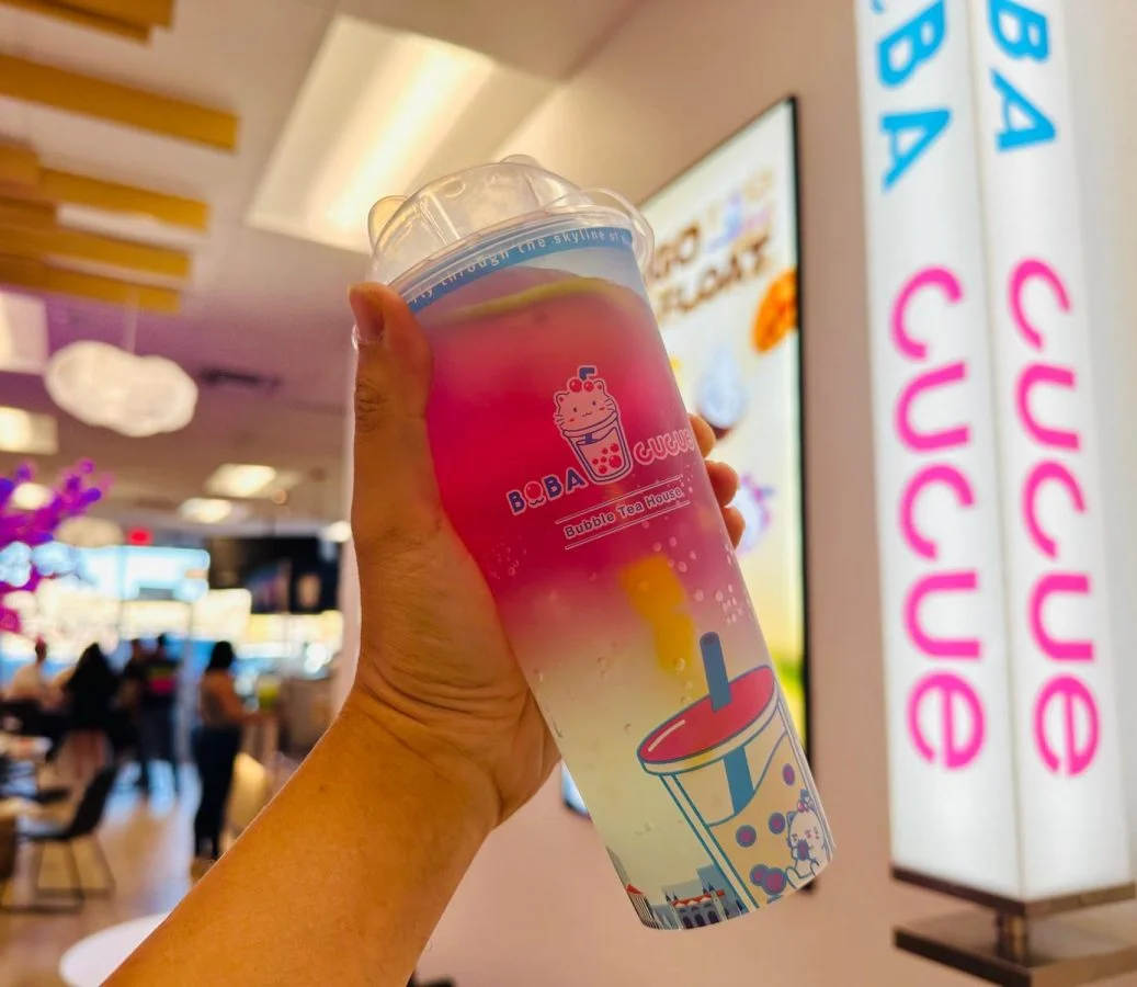 Hand holding gradient Boba Cucue bubble tea cup in the bubble tea shop interior.