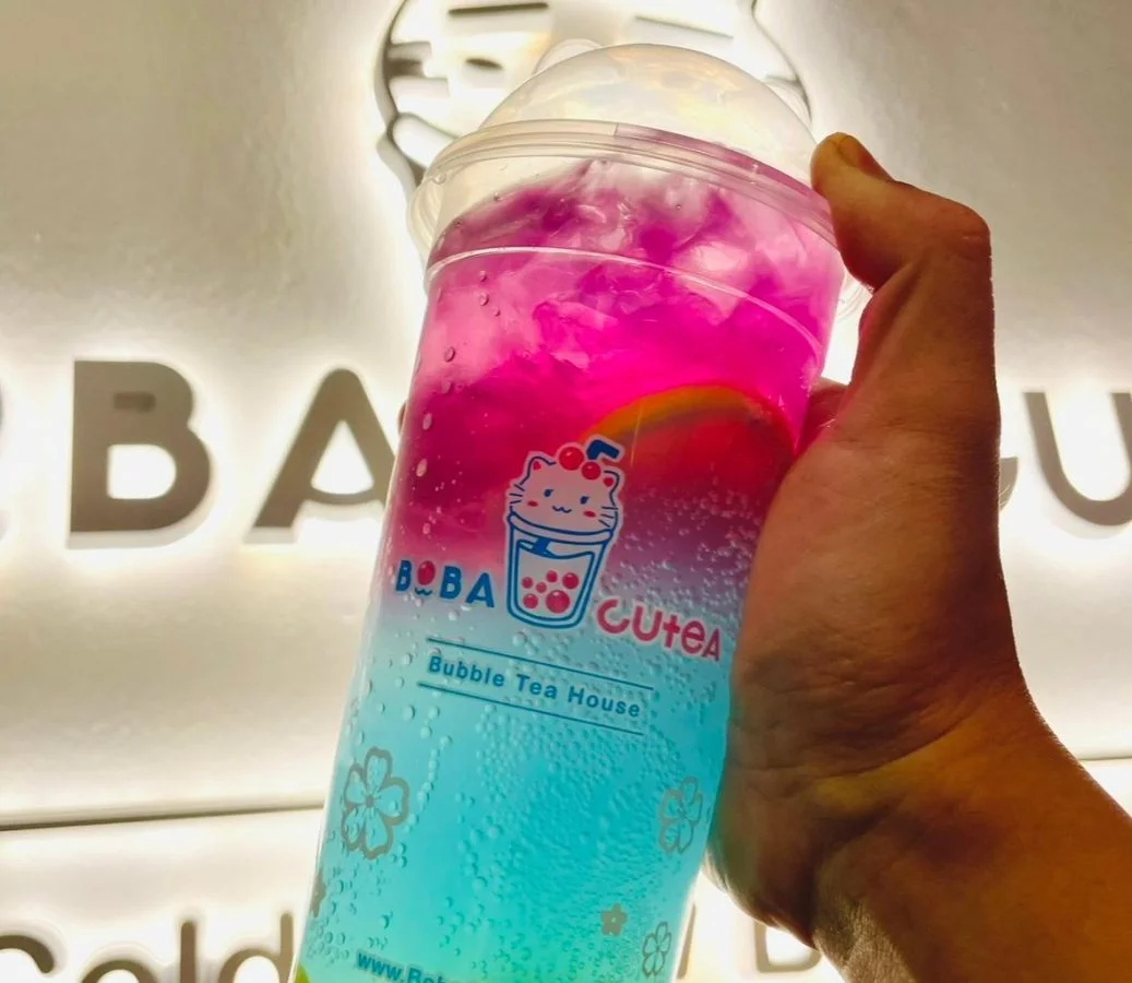 Hand holding colorful Boba Cutea bubble tea cup against branded shop wall.